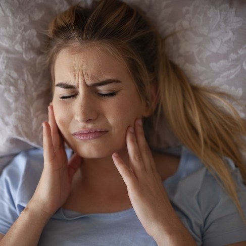 Woman laying in bed and wincing while holding her jaw in pain